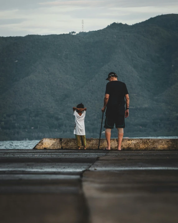 a man and a child standing on a pier, pexels contest winner, on the concrete ground, hills in the background, black sand, snapchat photo