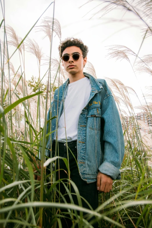 a man standing in a field of tall grass, an album cover, by Robbie Trevino, trending on unsplash, photorealism, jean jacket, casual pose, set on singaporean aesthetic, wear ray - ban glass