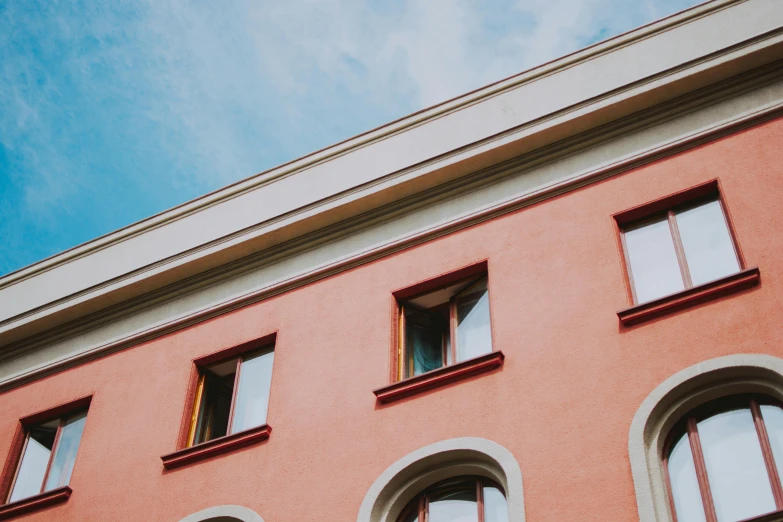 a red building with windows and a blue sky in the background, inspired by Wes Anderson, pexels contest winner, neoclassicism, faded colors, view from below, pink and orange, before a stucco wall