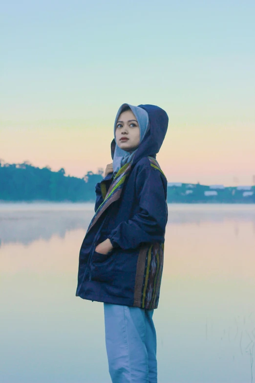 a woman standing in front of a body of water, a picture, inspired by Bernardino Mei, unsplash, realism, blue parka, hijab, 🤤 girl portrait, dusk