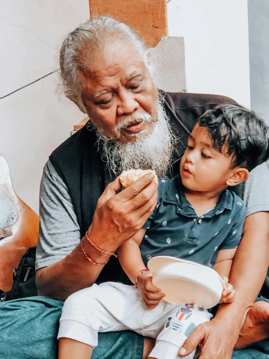 a man sitting on top of a couch holding a child, by Bapu, pexels contest winner, hurufiyya, eating cakes, white hair and white beard, profile image, mixed race