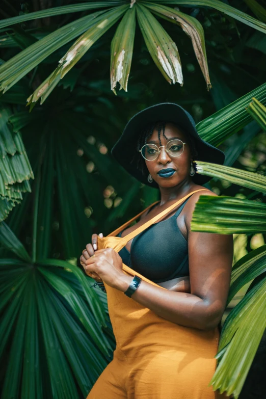 a woman standing in front of a palm tree, by Lily Delissa Joseph, pexels contest winner, afrofuturism, wearing black camisole outfit, dark green leaves, wearing black rimmed glasses, richly colored