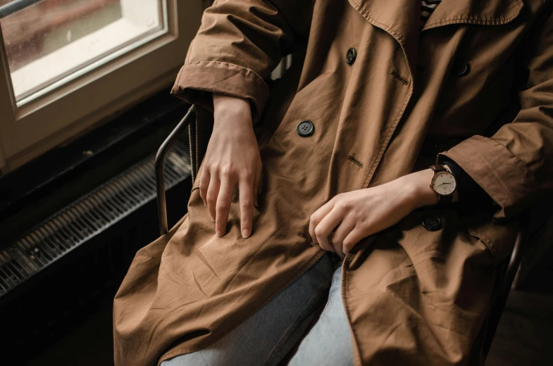 a woman sitting in a chair next to a window, trending on pexels, light brown trenchcoat, high angle close up shot, wearing a watch, sustainable materials