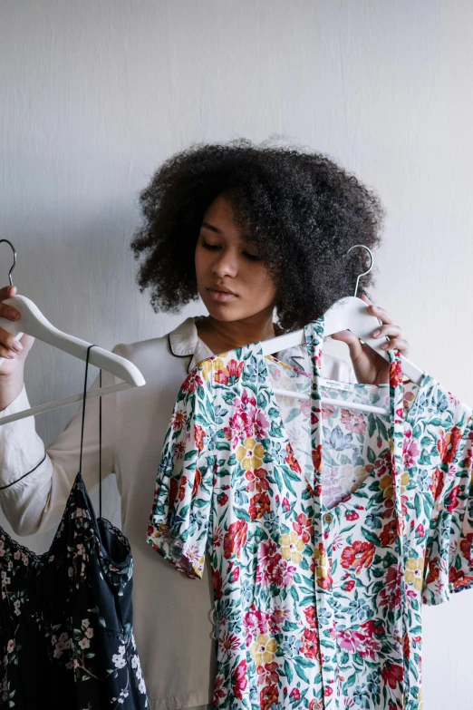 a woman standing next to a rack of clothes, flowery, with afro, wearing a white button up shirt, looking her shoulder