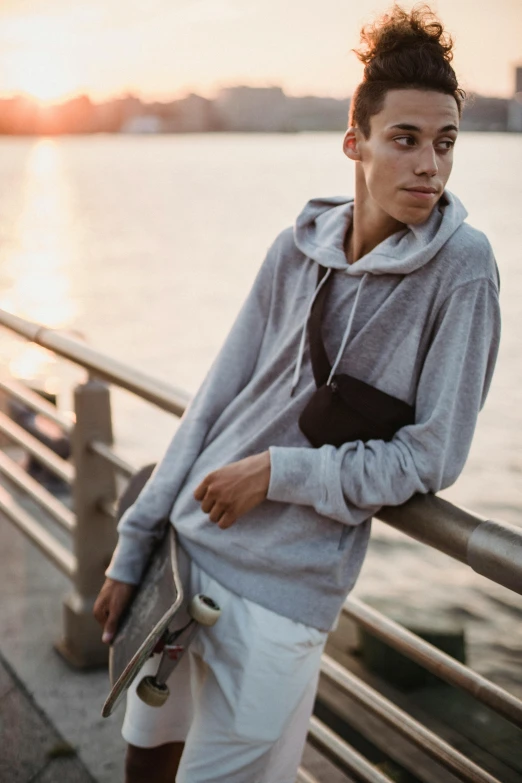 a man standing next to a body of water holding a skateboard, grey hoodie, pouches, summer evening, boy with neutral face