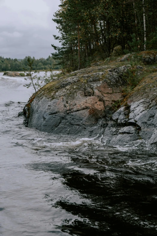 a man riding a surfboard on top of a river, inspired by Eero Järnefelt, unsplash, hurufiyya, with lots of dark grey rocks, panorama shot, trees bent over river, overcast mood