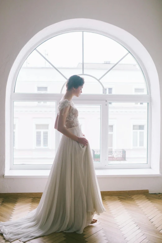 a woman in a wedding dress standing in front of a window, inspired by Antoni Brodowski, pexels contest winner, big arches in the back, inside white room, style of julia razumova, 3 / 4 view portrait