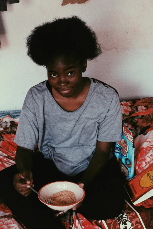 a person sitting on a bed with a bowl of food, by Lily Delissa Joseph, teenager, dark skinned, cereal, crisp face