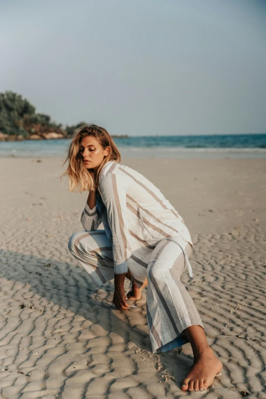 a woman kneeling in the sand on a beach, by Alice Mason, trending on unsplash, wearing stripe shirt, wearing white pajamas, fashion model features, girl in suit