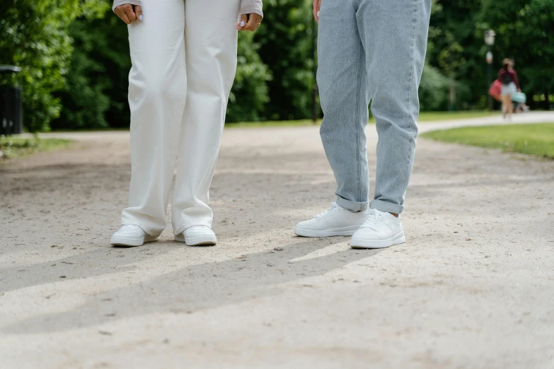 a man and a woman standing next to each other, trending on pexels, wearing white sneakers, parks and public space, background image, white pants