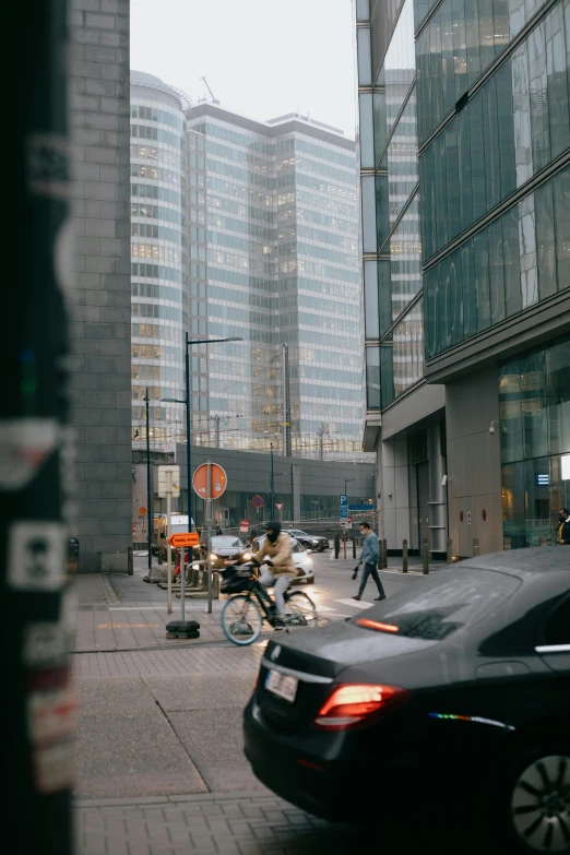 a black car driving down a street next to tall buildings, by Patrick Ching, pexels contest winner, hazy and dreary, people walking around, riding a motorbike down a street, buildings made out of glass