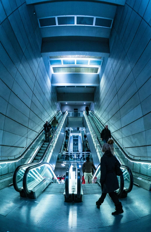 a group of people riding down an escalator, warsaw, in the evening, underground facility, light blues