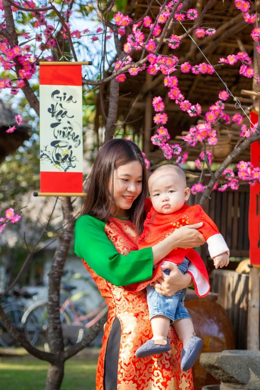 a woman holding a baby in her arms, a picture, inspired by Cui Bai, pexels contest winner, red flags holiday, wearing authentic attire, square, blossoms