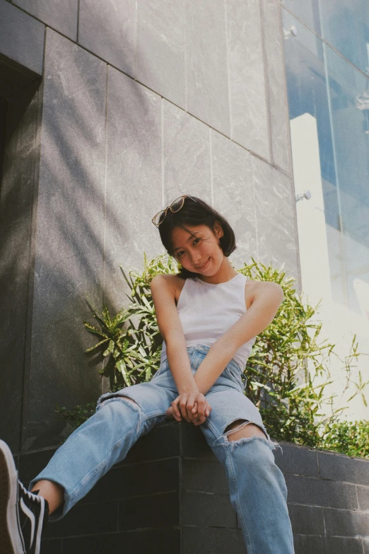 a woman sitting on a ledge in front of a building, by Tan Ting-pho, pexels contest winner, outfit : jeans and white vest, young cute wan asian face, sunfaded, smiling down from above