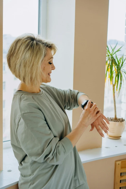 a woman standing in front of a window next to a potted plant, heart rate, sitting with wrists together, connectivity, blond