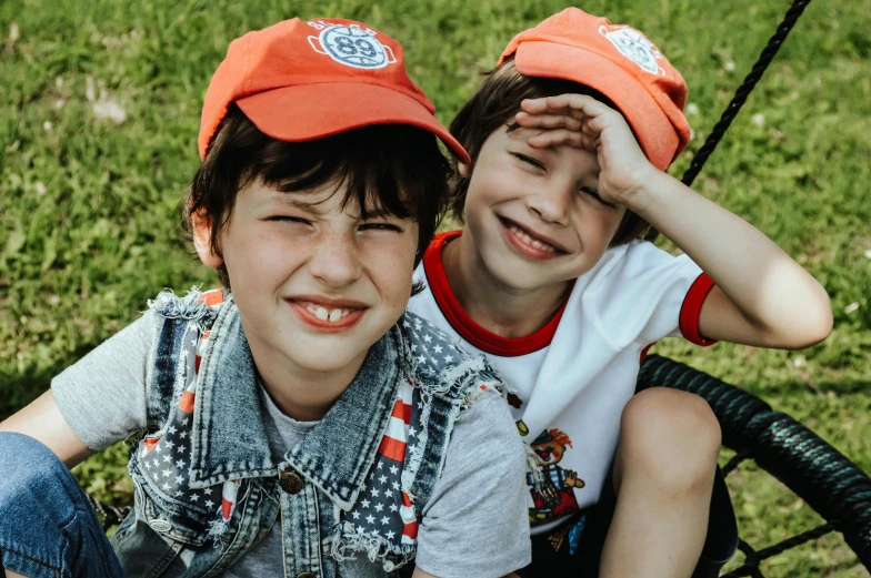 a couple of young boys sitting next to each other, by Lucia Peka, pexels contest winner, red cap, round teeth and goofy face, lookbook, postman pat