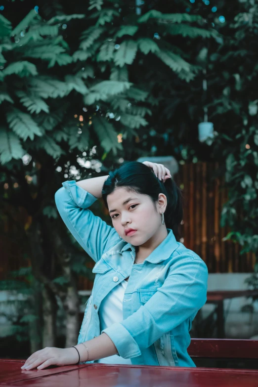 a woman sitting at a table with her hand on her head, inspired by Elsa Bleda, unsplash, wearing a jeans jackets, south east asian with round face, 15081959 21121991 01012000 4k, 🤤 girl portrait