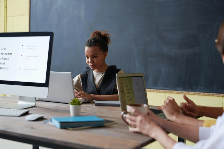 a group of people sitting at a table in front of a computer, blackboard in background, alana fletcher, getty images, standing in class