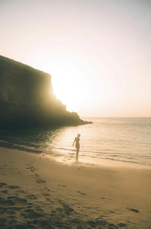 a person standing on top of a beach next to the ocean, sun lit, cliffs, sunset beach, white sandy beach