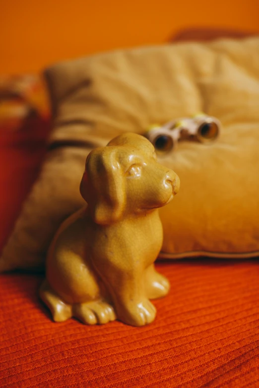 a dog figurine sitting on a bed next to a pillow, inspired by Sarah Lucas, shades of aerochrome gold, in claymation, medium close up, soap