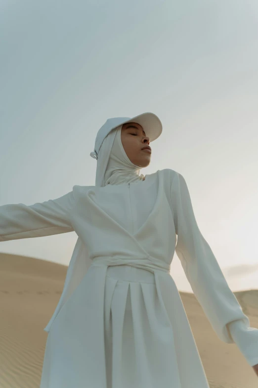 a woman in a white dress standing in the desert, an album cover, inspired by Amir Zand, trending on pexels, hurufiyya, hat and hoodie, white suit, portrait mode photo, afternoon