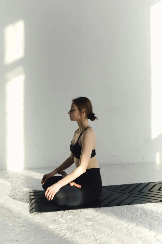 a woman sitting on a mat in a room, inspired by Fei Danxu, unsplash, light and space, wearing a cropped black tank top, gif, sukhasana, low quality photo