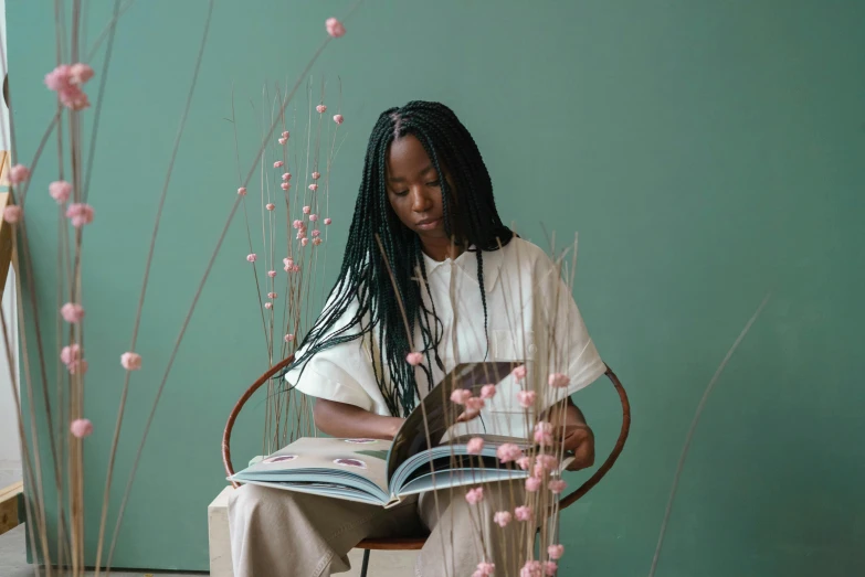 a woman sitting in a chair reading a book, by Lily Delissa Joseph, pexels contest winner, black arts movement, branches growing as hair, adut akech, sea - green and white clothes, brown and pink color scheme