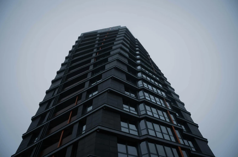 a tall black building with lots of windows, by Emma Andijewska, unsplash, under a gray foggy sky, ten flats, dark grey and orange colours, view from bottom to top