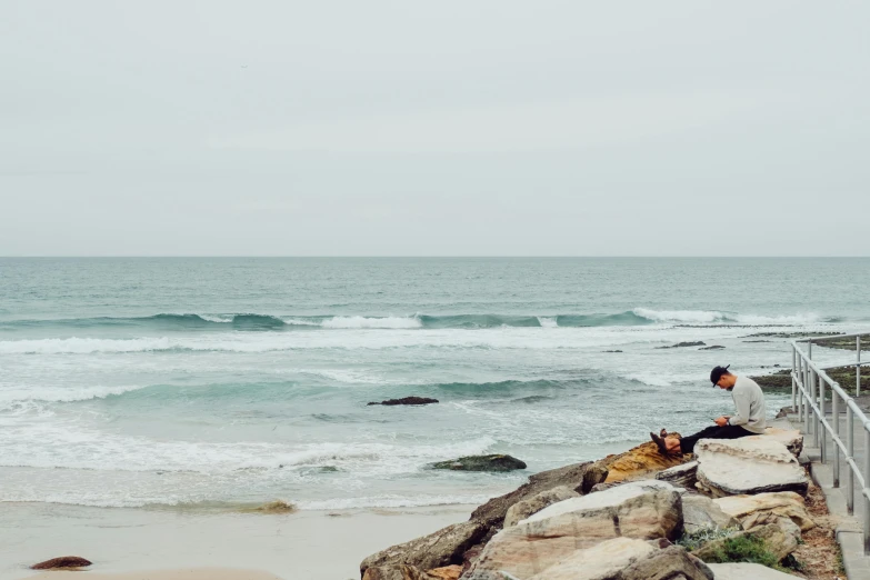 a man sitting on top of a rock next to the ocean, by Nina Hamnett, pexels contest winner, australian beach, slight overcast, surfing, panoramic view of girl