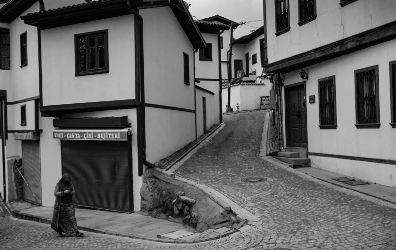 a black and white photo of a woman walking down a cobblestone street, by Tamas Galambos, built on a steep hill, wooden houses, gui guimaraes, white wall complex