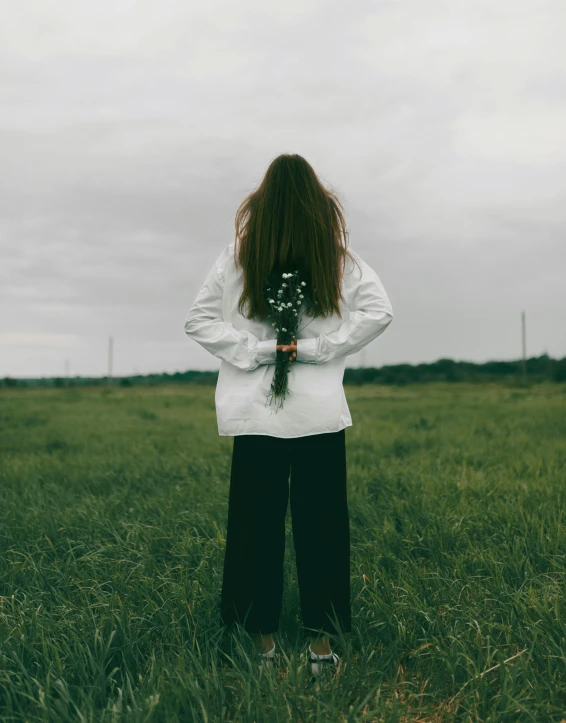 a woman standing in a field holding a bouquet of flowers, an album cover, by Attila Meszlenyi, trending on unsplash, queer woman, wearing a white sweater, loneliness, unsettling image