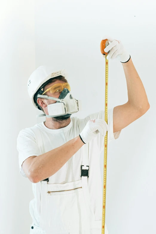 a man in white overalls holding a tape measure, pexels contest winner, dust mask, helmet on, tall ceiling, plain background