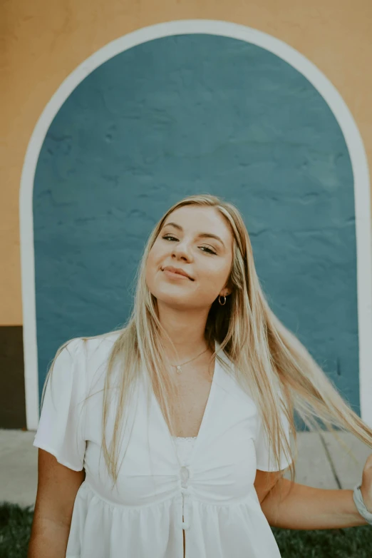 a woman standing in front of a blue and yellow building, a black and white photo, by Olivia Peguero, trending on unsplash, long blonde hair and blue eyes, smiling young woman, in front of white back drop, beautifully lit