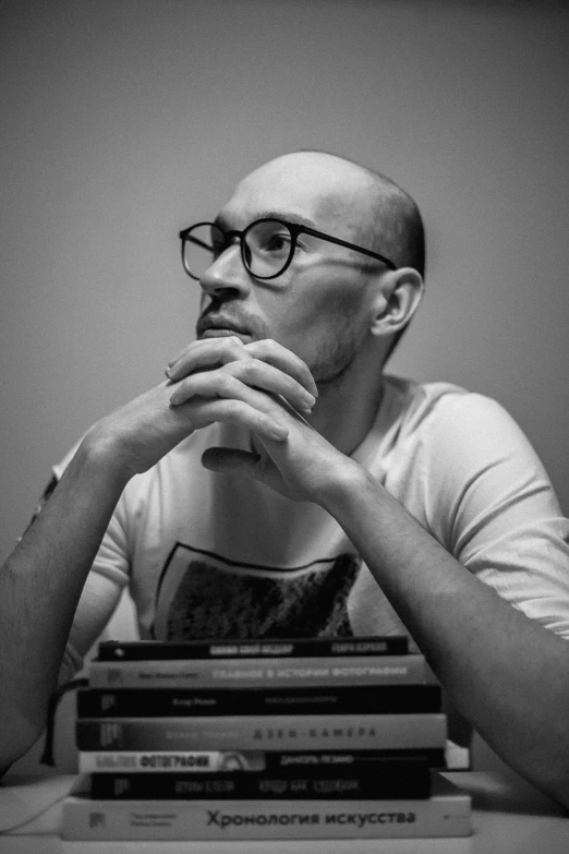 a man sitting at a table with a stack of books, a black and white photo, inspired by Leo Leuppi, hyperrealism, bald with short beard, twitch streamer / gamer ludwig, vincenzo riccardi, man with glasses