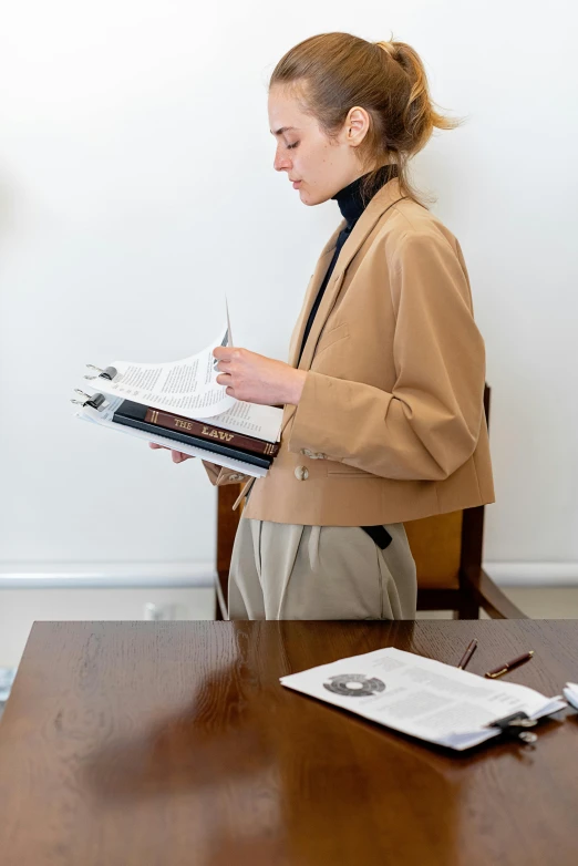 a woman sitting at a table writing on a piece of paper, by Nina Hamnett, private press, light brown coat, lawyer clothing, holding a giant book, trending photo