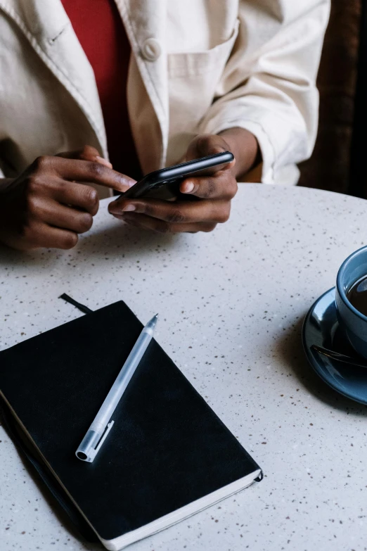 a person sitting at a table using a cell phone, trending on pexels, sitting on a mocha-colored table, black, scholarly, dark-skinned