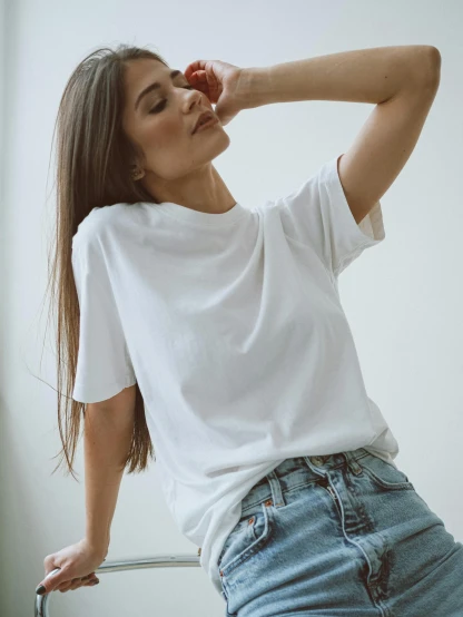 a woman sitting on top of a chair next to a window, by Robbie Trevino, trending on unsplash, white t-shirt, model standing pose, detailed product image, slightly rounded face