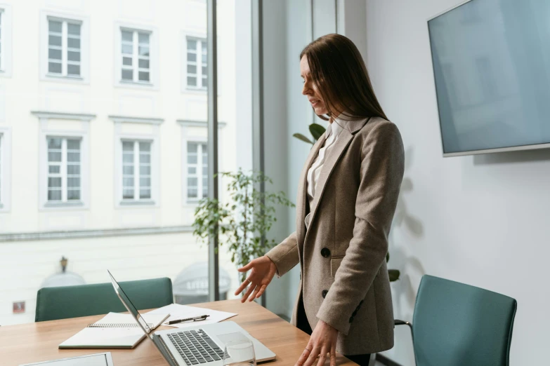 a woman standing in front of a laptop computer, by Emma Andijewska, pexels contest winner, in a meeting room, bend over posture, girl in a suit, noelle stevenson