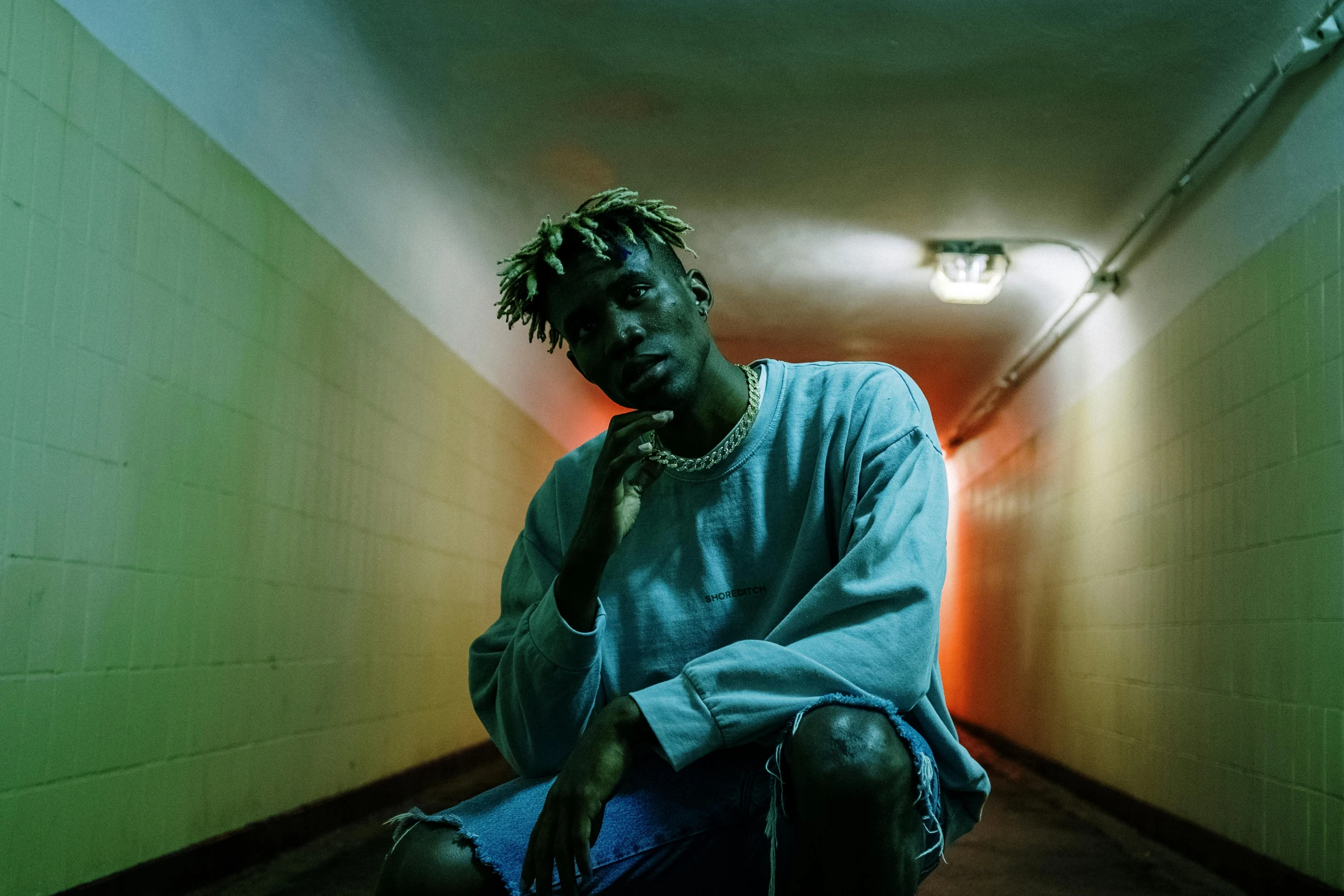 a man sitting on the ground in a hallway, an album cover, trending on pexels, visual art, xxxtentacion, hero pose colorful city lighting, with teal clothes, profile image