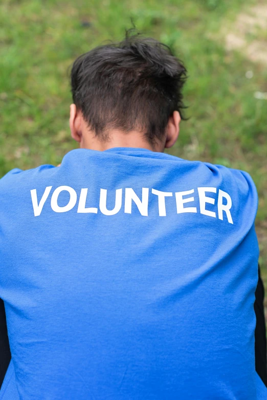 a man sitting on a bench wearing a volunteer t - shirt, pexels contest winner, happening, showing her shoulder from back, blue uniform, standing in a grassy field, promotional image