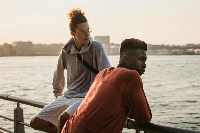 two men sitting next to each other near a body of water, trending on pexels, happening, afropunk, looking her shoulder, androgynous person, lightweight