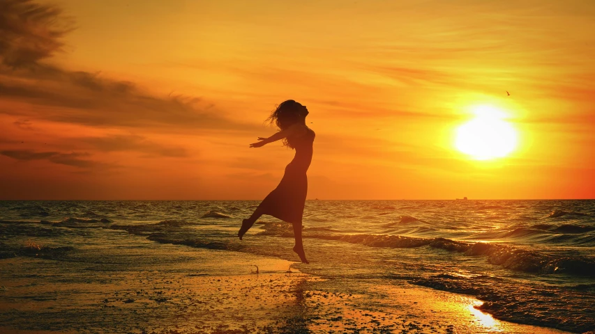 a woman standing on top of a beach next to the ocean, pexels contest winner, romanticism, flying through sunset, yellow, leaping, lynn skordal