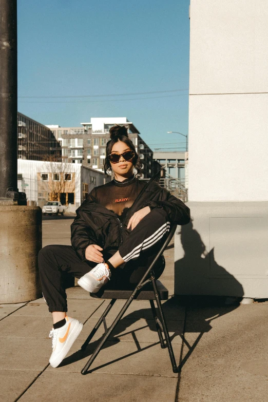 a woman sitting in a chair on a sidewalk, an album cover, trending on unsplash, wearing adidas clothing, shady look, casual black clothing, with sunglass