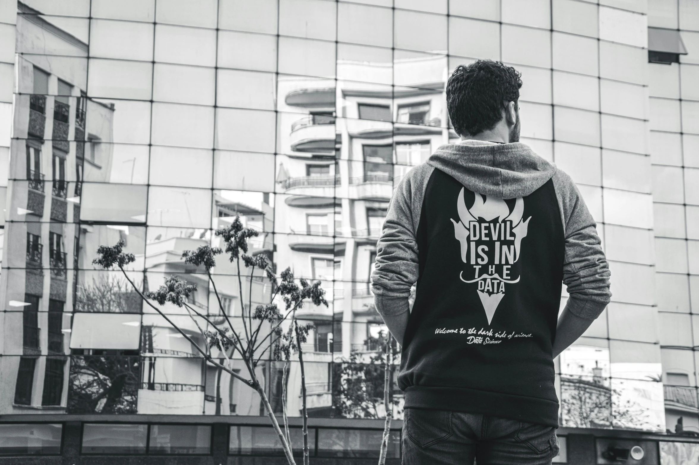 a man standing in front of a tall building, a black and white photo, inspired by Anton Ažbe, pexels contest winner, graffiti, hoodie, skull image on the vest, her back is to us, in style of dark souls 3