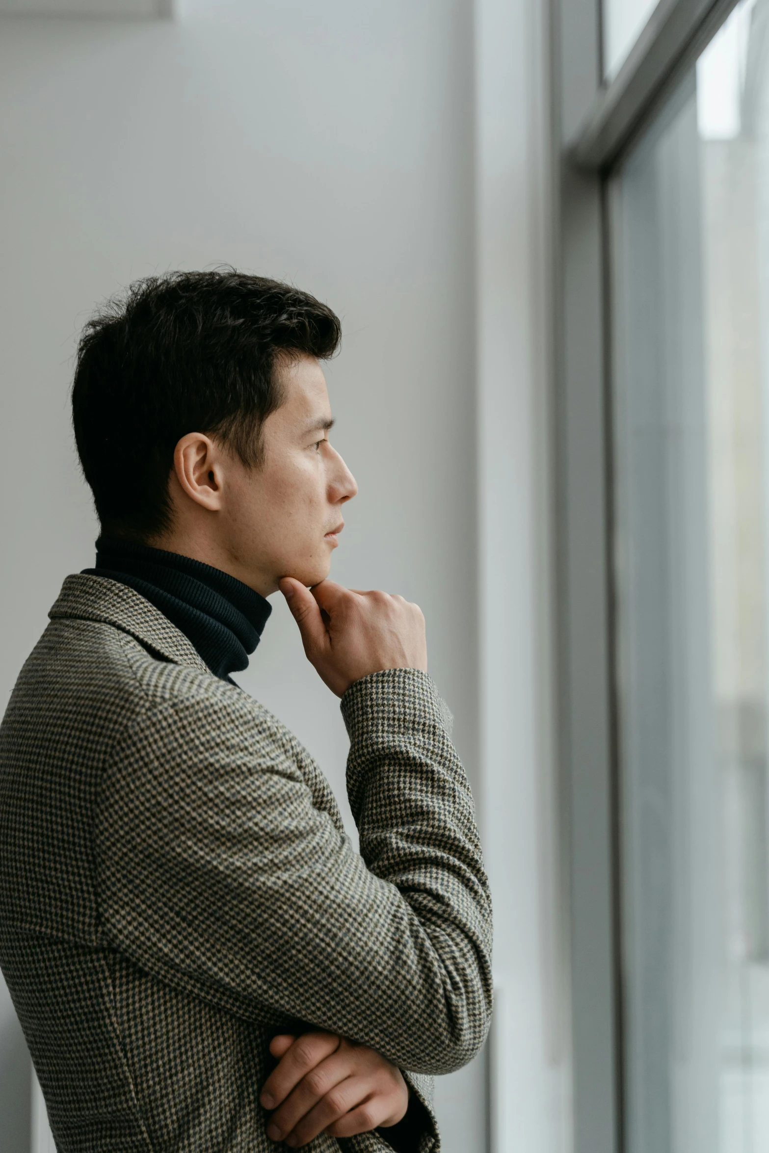 a man standing in front of a window with his hand on his chin, inspired by Fei Danxu, trending on pexels, grey turtleneck coat, professional profile picture, androgynous person, facing away