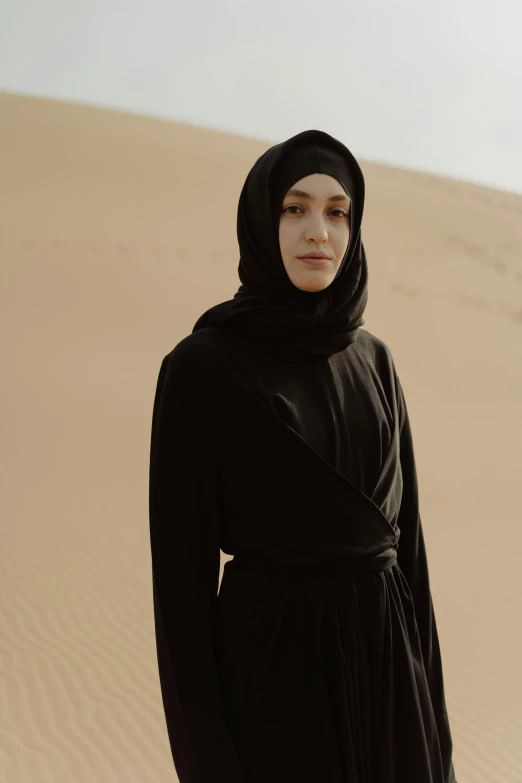 a woman in a black dress standing in the desert, inspired by Maryam Hashemi, hurufiyya, main colour - black, looking serious, official product photo, mecca