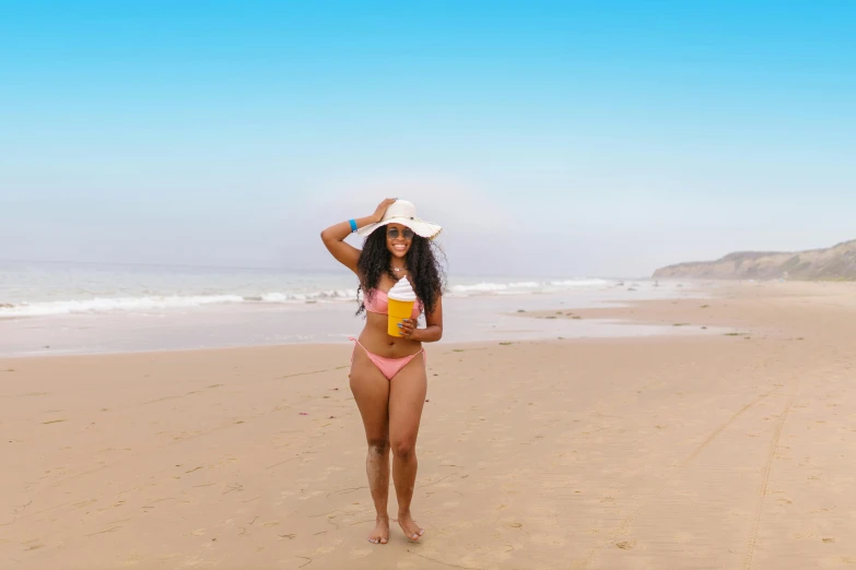 a woman standing on top of a sandy beach, by Lily Delissa Joseph, pexels contest winner, holding a drink, curvy body, ( brown skin ), with hat