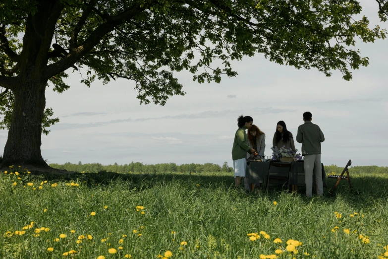 a group of people sitting around a table under a tree, a picture, unsplash, land art, standing in a field with flowers, having a picnic, conde nast traveler photo