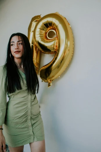 a woman standing in front of a gold number 9 balloon, pexels contest winner, young woman with long dark hair, tight wrinkled cloath, profile image, indoor picture