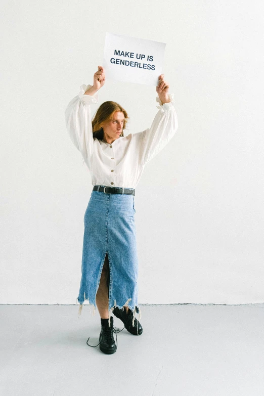 a woman holding up a sign in front of a white wall, an album cover, trending on unsplash, private press, denim, dressed in long fluent skirt, wearing business casual dress, kindness